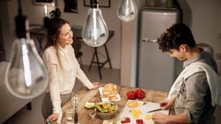 Une femme observe un homme couper des légumes sur une planche à découper dans une cuisine ; on aperçoit des ampoules dans la partie supérieure de l’image.